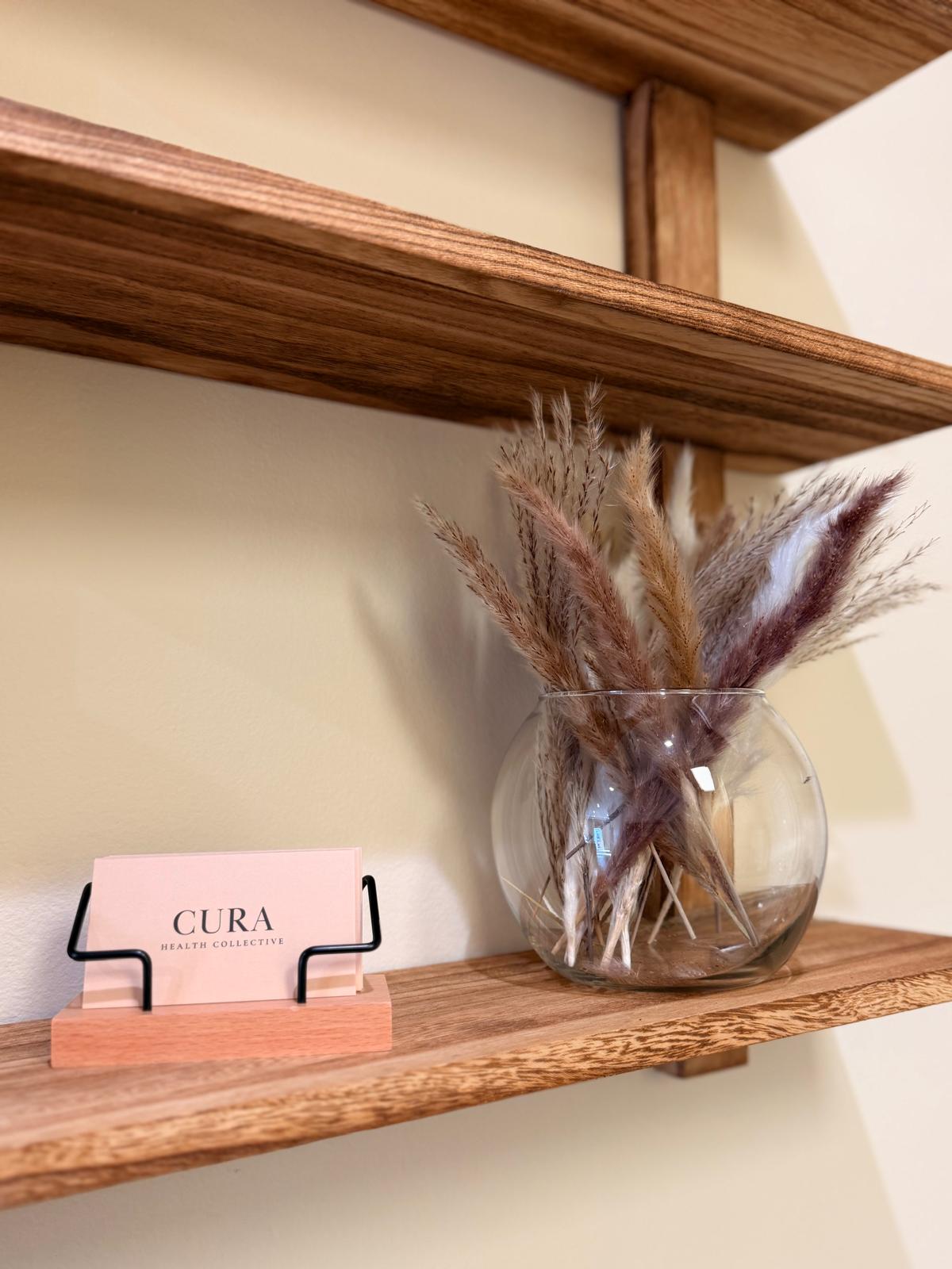 A Cura Health Collective business card on a wooden shelf beside dried pampas grass